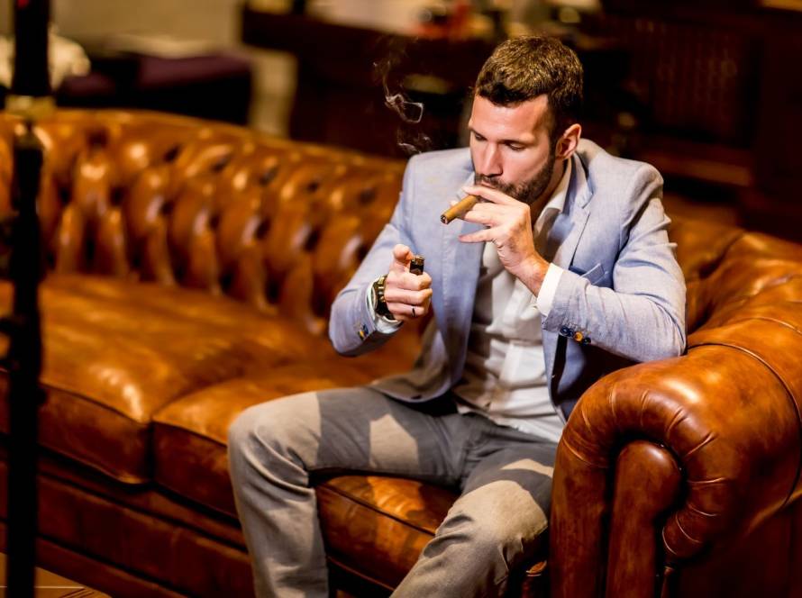 Young businessman in a tailored suit lighting a cigar while seated on a leather couch, with a glass of white wine resting on the table.