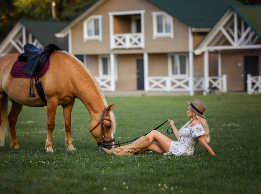 A woman wearing a floral dress and a cowboy hat sits on the grass while holding the horse’s leash as it grazes