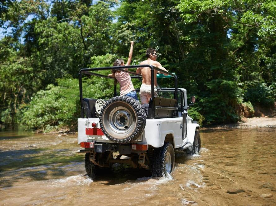 Young couple standing at the back of an off-road vehicle while crossing a shallow river during an excursion